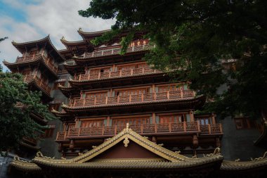 Geleneksel Çin mimarisinde koyu gri duvarları ve yemyeşil çam ağaçlarıyla çevrili kırmızı-kahverengi ahşap aksanları olan çok katmanlı bir bina var..