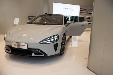 Açık gri Xiaomi SU7 elektrikli sedan açık sürücü kapısı parlak modern sergi salonunun beyaz cilalı zemininde duruyor..