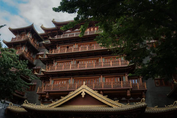 Geleneksel Çin mimarisinde koyu gri duvarları ve yemyeşil çam ağaçlarıyla çevrili kırmızı-kahverengi ahşap aksanları olan çok katmanlı bir bina var..