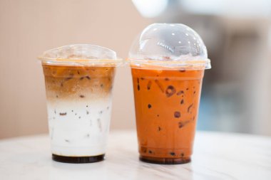 İki şeffaf plastik bardakta beyaz mermer bir masanın üzerinde oturuyor, biri katmanlı buzlu kahve, diğeri ise turuncu renkli buzlu Tayland çayı içeriyor..