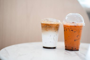 İki şeffaf plastik bardakta beyaz mermer bir masanın üzerinde oturuyor, biri katmanlı buzlu kahve, diğeri ise turuncu renkli buzlu Tayland çayı içeriyor..