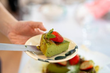 Bir dilim yeşil kibrit peynirli kek ile küçük bir tabak tutarken üstüne taze çilek ve nane yaprağı konmuş..