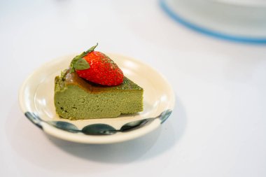 Üçgen dilimli yeşil kibrit peynirli kek üzerine taze çilek konmuş yarısı parlak beyaz arka planda küçük beyaz seramik tabakta duruyor..