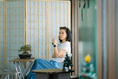 Asyalı bir kadın, parlak bir odada, yeşil bir içecekle küçük tahta bir masada oturuyor ve kameraya bakıyor.