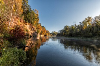 Sigulda, Letonya 'daki Gauja nehri üzerindeki kırmızı kumtaşı kayalıklarının güneşli bir sonbahar günü manzarası.