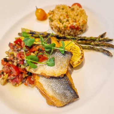 A vibrant dish of crispy-skinned seabass fish fillets served with a vibrant tomato and olive salsa relish, stalks of grilled asparagus and a scoop of rice pilaf topped with a grilled lemon slice