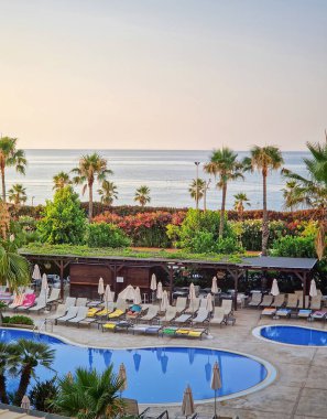 Günbatımında sakin tropik tatil köyü manzarası. Ön planda geniş, mavi bir yüzme havuzu. Etrafı şezlonglar ve palmiye ağaçlarıyla çevrili. Yumuşak, pastel bir gökyüzünün altında ufka uzanan sakin bir okyanus.