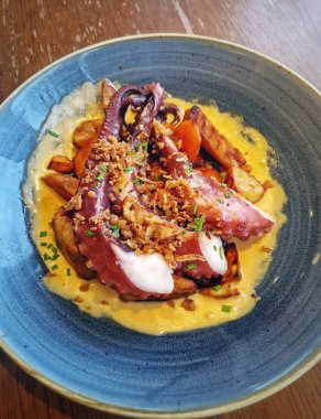 Beautifully plated dish of tender grilled octopus tentacles served over golden roasted sweet potatoes and creamy sauce, garnished with crispy fried onions, presenting a gourmet culinary experienc