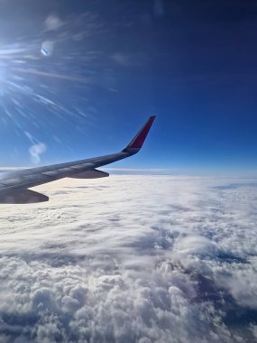 Bulutların üstünde uçak uçuşu. Mavi gökyüzü ve uçağın kanadı pencereden görülüyor.