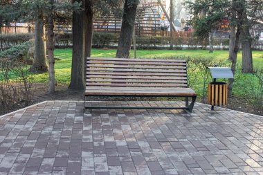 Parlak siyah metal çerçeveli ahşap bir bankta, yumuşak bir sabah ışığı altında, birbirine uyumlu modern bir çöp kutusu eşliğinde, sakin, ağaçlarla kaplı bir parkta, çok tonlu bir tuğla patikada oturuyor..
