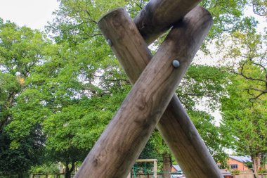 Kalın kütüklerden yapılmış doğal bir ahşap tırmanma iskeletinin yakın çekimi, sürgülü eklemler içeriyor. Yapı, yemyeşil ağaçların ve bir yerleşim yerinin arka planına karşı dışarıda duruyor..