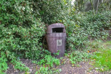 Kahverengi, ahşap bir çöp tenekesi, kalın yeşil çalılıklarda gömülü duruyor. Kapüşonlu kaputu yağmur girişini engelliyor ve yan tarafındaki gümüş simge çöp boşaltan kişiyi gösteriyor.