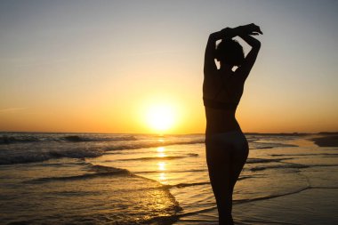 Denizin üzerindeki altın gün batımında kumsalda poz veren zayıf kadın silueti. Gündoğumunu ve günbatımını takip ederek aktif güzellik yaşam tarzı, özgürlük kavramı