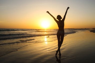 Denizin üzerindeki altın gün batımında kumsalda poz veren zayıf kadın silueti. Gündoğumunu ve günbatımını takip ederek aktif güzellik yaşam tarzı, özgürlük kavramı