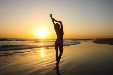 Denizin üzerindeki altın gün batımında kumsalda poz veren zayıf kadın silueti. Gündoğumunu ve günbatımını takip ederek aktif güzellik yaşam tarzı, özgürlük kavramı