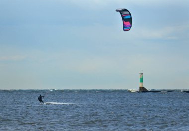 Bir uçurtma sörfçüsü bulutlu bir günde arka planda deniz feneriyle rüzgar sörfü yapıyor.