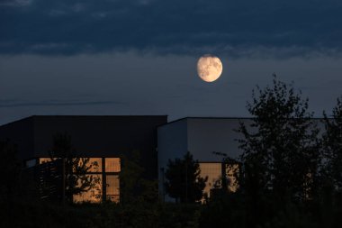 A night or twilight landscape with a full moon shining brightly in a dark, cloudy sky. Silhouettes of modern buildings are visible in the foreground.