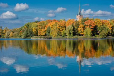 A landscape where the pointed spire and the top of an ancient tower rise above a dense line of autumn trees. The vibrant foliage is tinged with rich yellow and orange tones.
