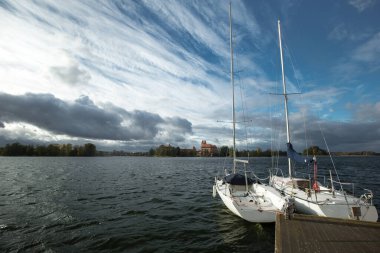 Trakai Kalesi 'nin, gölün ortasındaki bir adada çekilmiş manzarası. Ön planda, iki beyaz yelkenli yatı ahşap bir rıhtıma demirlemiş..