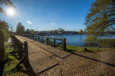 Güneşli bir sonbahar gününde çekilen geniş açılı çekim, bir körfez veya gölün bir bölümünü geçen uzun ve alçak bir ahşap köprüyü resmediyor. Ön planda düşmüş yapraklarla kaplı bir kaldırım taşı yolu var..