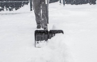 Gri elbiseli bir adam, özel bir evin bahçesinde siyah plastik bir kürekle yeni yağmış bir karı temizliyor. Kar fırtınası sırasında açık havada kış çalışması.