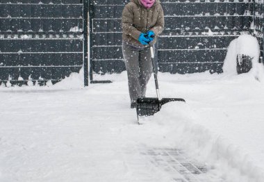 Gri elbiseli bir adam, özel bir evin bahçesinde siyah plastik bir kürekle yeni yağmış bir karı temizliyor. Kar fırtınası sırasında açık havada kış çalışması.