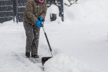 Gri elbiseli bir adam, özel bir evin bahçesinde siyah plastik bir kürekle yeni yağmış bir karı temizliyor. Kar fırtınası sırasında açık havada kış çalışması.