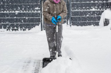 Gri elbiseli bir adam, özel bir evin bahçesinde siyah plastik bir kürekle yeni yağmış bir karı temizliyor. Kar fırtınası sırasında açık havada kış çalışması.