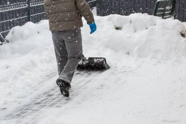 Gri elbiseli bir adam, özel bir evin bahçesinde siyah plastik bir kürekle yeni yağmış bir karı temizliyor. Kar fırtınası sırasında açık havada kış çalışması.