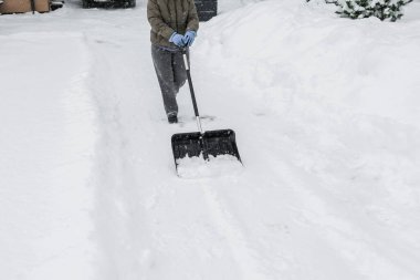 Gri elbiseli bir adam, özel bir evin bahçesinde siyah plastik bir kürekle yeni yağmış bir karı temizliyor. Kar fırtınası sırasında açık havada kış çalışması.