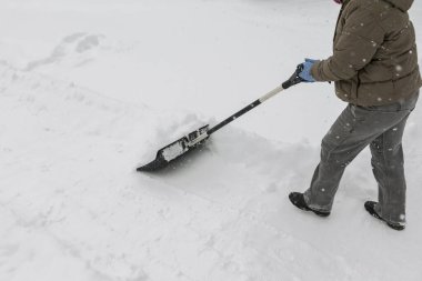 Gri elbiseli bir adam, özel bir evin bahçesinde siyah plastik bir kürekle yeni yağmış bir karı temizliyor. Kar fırtınası sırasında açık havada kış çalışması.