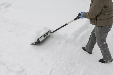 Gri elbiseli bir adam, özel bir evin bahçesinde siyah plastik bir kürekle yeni yağmış bir karı temizliyor. Kar fırtınası sırasında açık havada kış çalışması.
