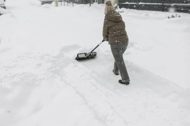 Gri elbiseli bir adam, özel bir evin bahçesinde siyah plastik bir kürekle yeni yağmış bir karı temizliyor. Kar fırtınası sırasında açık havada kış çalışması.
