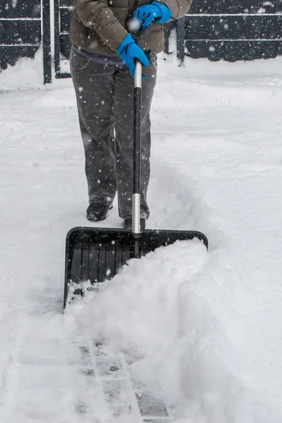 Gri elbiseli bir adam, özel bir evin bahçesinde siyah plastik bir kürekle yeni yağmış bir karı temizliyor. Kar fırtınası sırasında açık havada kış çalışması.