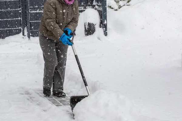 Gri elbiseli bir adam, özel bir evin bahçesinde siyah plastik bir kürekle yeni yağmış bir karı temizliyor. Kar fırtınası sırasında açık havada kış çalışması.