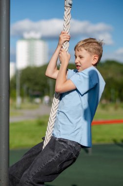 Genç çocuk kendini halatla yukarı çekiyor. Spor sahası, eğitim anlayışı, güç, dayanıklılık ve aktif yaşam tarzı için çabalıyor.