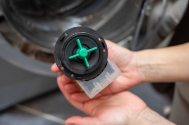 Çamaşır makinesi pompası filtresi tutan kişinin yakın plan fotoğrafı. Yeşil pervaneli, ev aletlerinin onarımı, temizliği ve bakımını gösteren ve yedek parçaları olan.