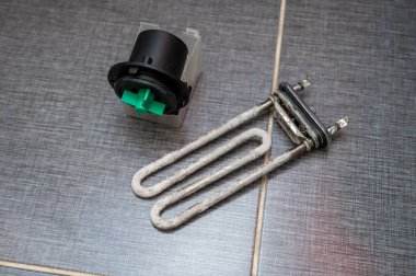 Used washing machine heating element covered with limescale and a pump motor with green impeller lie on a tiled surface, illustrating appliance maintenance or repair process