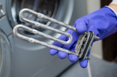 Mor temizlik eldiveni giyen kişi açık kapının yanında kireçle kaplanmış kullanılmış bir çamaşır makinesi ısıtma elementi tutuyor. Bu da parça değişimi ya da bakım sürecini gösteriyor.