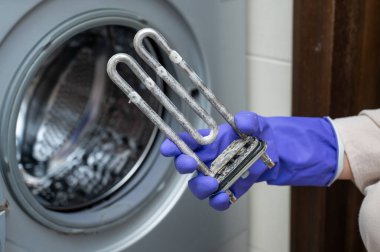 Mor temizlik eldiveni giyen kişi açık kapının yanında kireçle kaplanmış kullanılmış bir çamaşır makinesi ısıtma elementi tutuyor. Bu da parça değişimi ya da bakım sürecini gösteriyor.