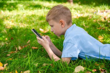 Genç bir çocuk yeşil çimlere uzanmış, akıllı telefonuna odaklanmış, alet bağımlılığını resmediyor.