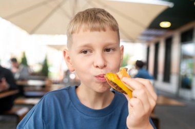 Genç sarışın çocuk açık hava kafede hamburgerle vakit geçiriyor. Huzurlu bir yaz günü. Normal ve dostane bir atmosfer.