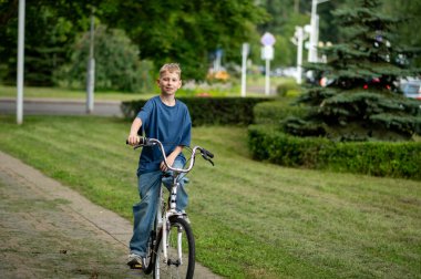 Sıcak bir yaz gününde yeşil parkta bisiklet süren neşeli sarışın çocuk, doğanın, özgürlüğün ve açık havada eğlencenin tadını çıkarıyor.