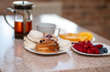 Konforlu mutfak sahnesi çaydanlık, marşmelov, taze çörekler ve meyveler tezgahın üstünde, sıcak ev havası.