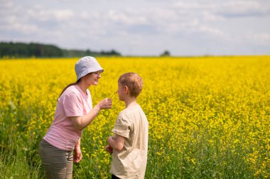 Anne, parlak sarı bir kolza tohumu tarlasında yan yana dururken oğluna kolza tohumu verir.