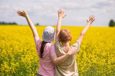 Sarı bir çiçek tarlasında dikilen iki mutlu çocuk, kollarını kaldırmış, yaz özgürlüğünün ve doğanın tadını çıkarıyorlar.