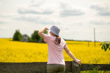 Genç bir kadın, alnında eliyle gözlerini koruyor. Çiçek açan bir kolza tohumu tarlasına ve kırsal bir tarlaya bakıyor.