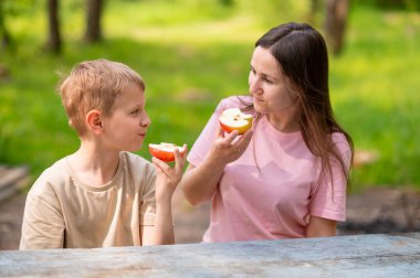 Anne ve oğul elma yiyor, gülümsüyor ve parkta meyve paylaşıyor.