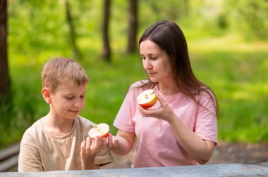 Anne ve oğul elma yiyor, gülümsüyor ve parkta meyve paylaşıyor.