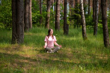 Huzurlu bir ormanda tek başına meditasyon yapan genç bir kadın, yeşil çimlerin üzerinde bağdaş kurarak oturuyor.